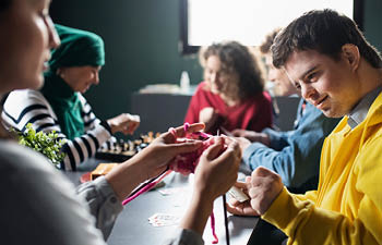 Direct Support Professionals and their clients gathered around a table, knitting and playing chess.