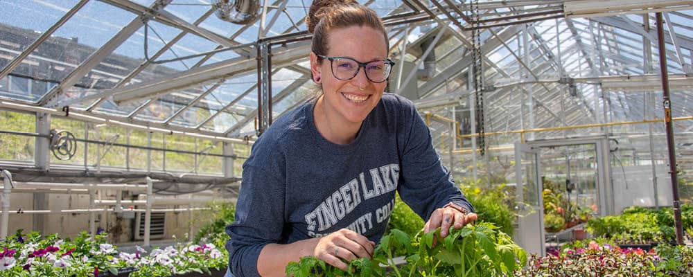 A young woman tending to her plants in the Finger Lakes Community College greenhouse pauses to smile at the camera.