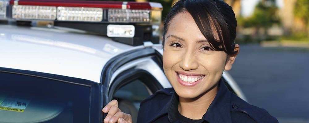 A police officer leaning against the side of her patrol car. She smiles and looks directly at the camera.