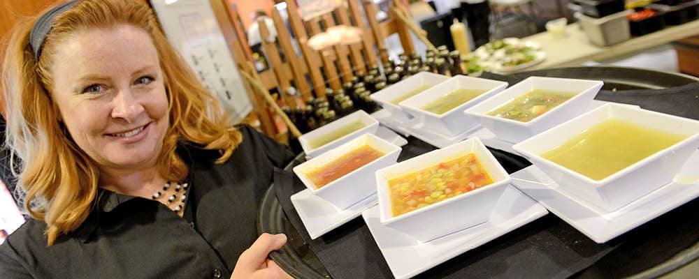 A server smiles at the camera while holding a food tray loaded with several bowls of soup prepared by FLCC Culinary Arts students.