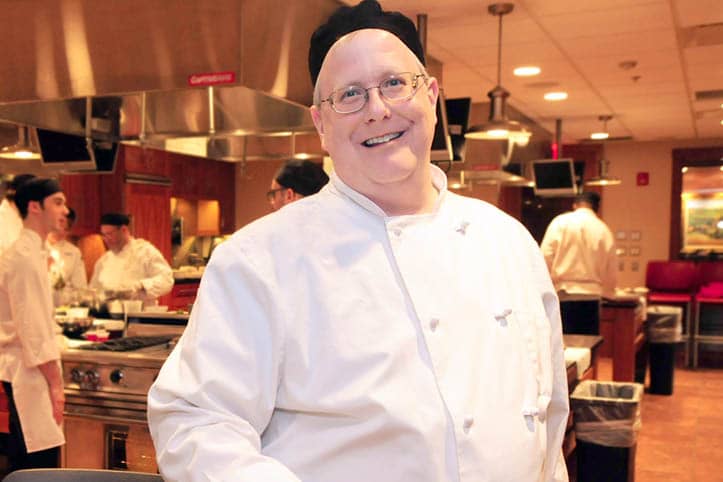 Culinary Arts faculty member poses for a picture. In the background, several Culinary Arts students are preparing food in a professional kitchen.