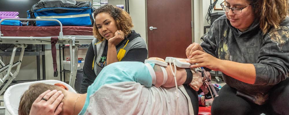 Two EMT students practice securing medical monitoring devices to a third student's arm. The volunteer patient is lying on his side as though injured.