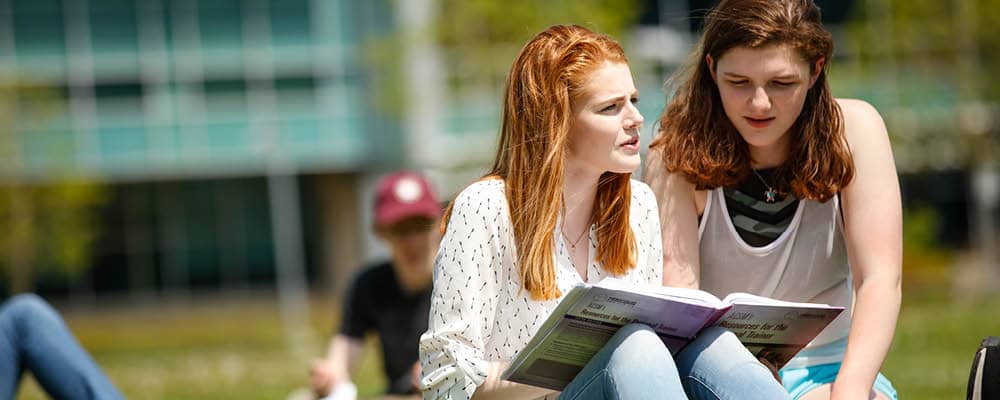 Two young women read from a college textbook together while lounging in the grass on a bright and sunny campus lawn.
